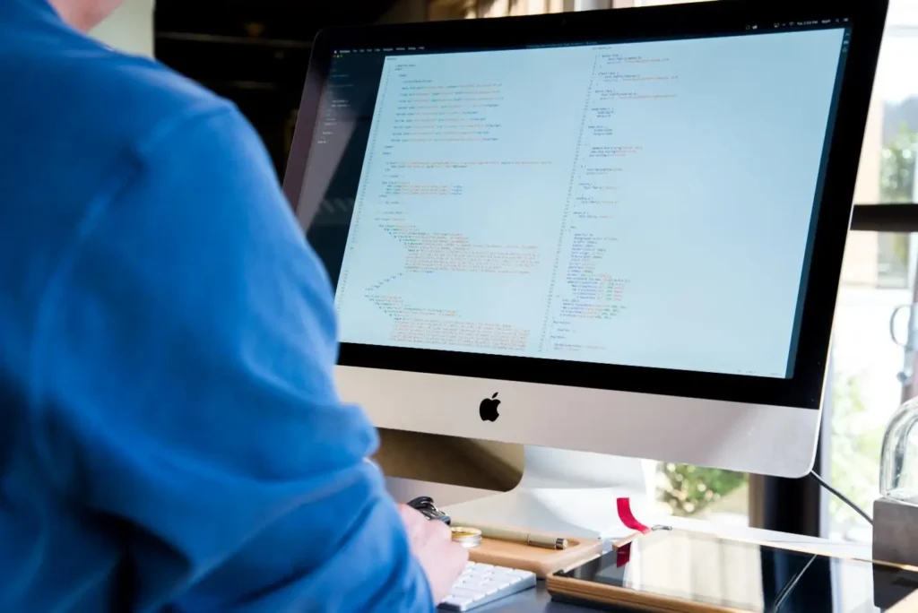 Person in a blue sweater viewed from behind, looking at an iMac displaying colorful code on screen.
